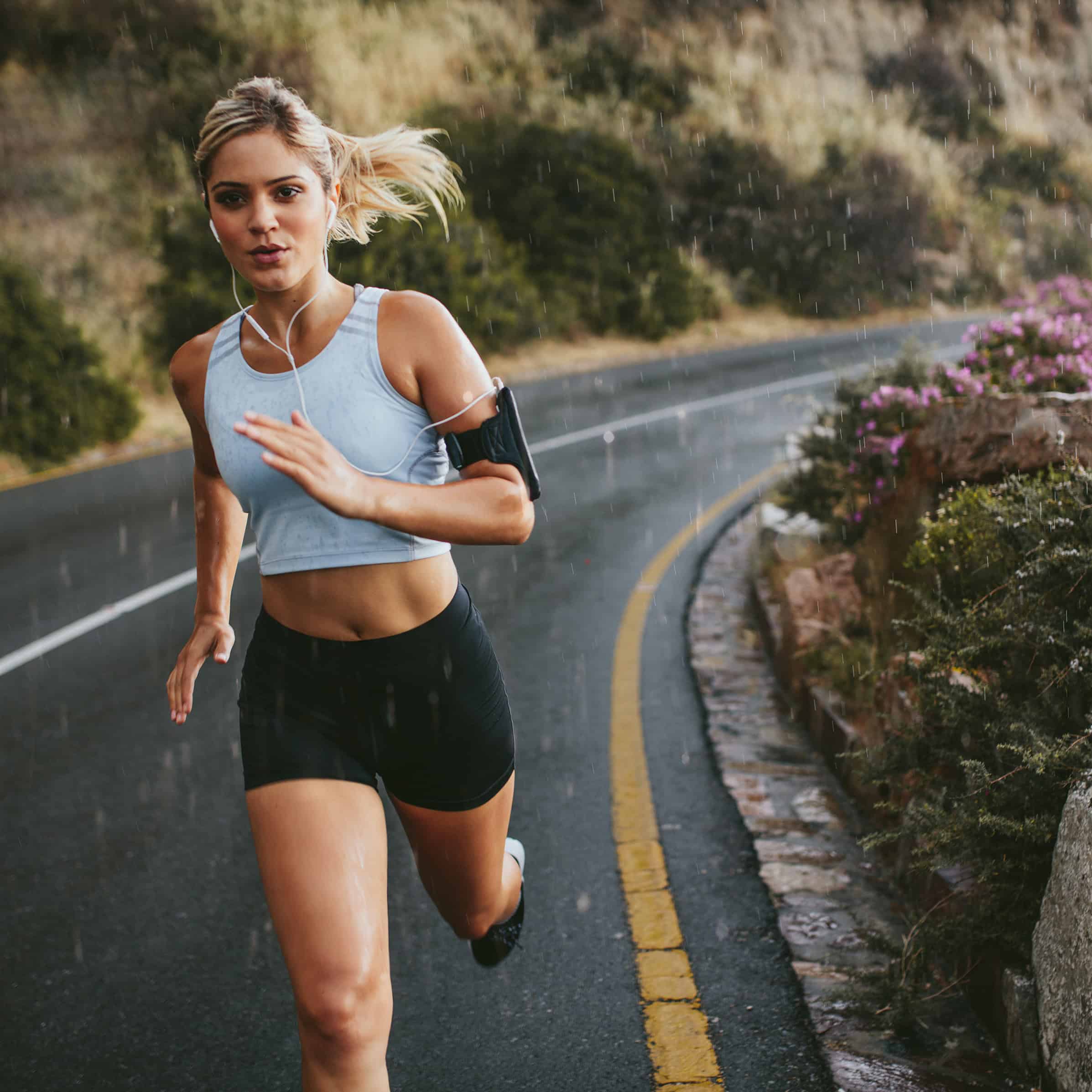 A determined female runner navigating a winding road in the rain on Chapman's Peak Drive, South Africa. Her commitment to peak physical performance is supported by the daily use of IMUMI Bovine Colostrum Powder, known for enhancing stamina and recovery.